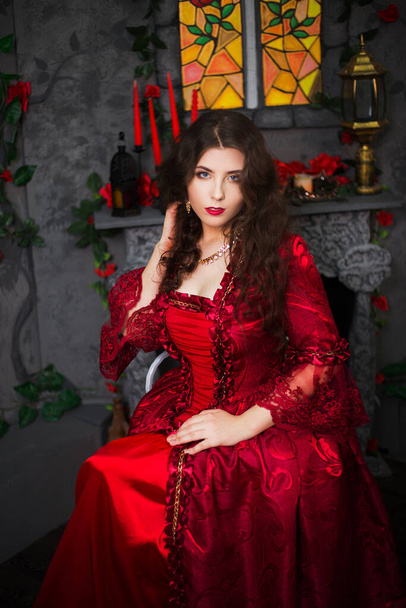 A beautiful girl in a magnificent red dress of the Rococo era sits on a chair near the fireplace, window and flowers. - Zdjęcie, obraz