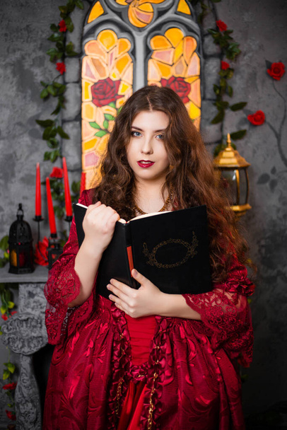 A beautiful girl in a magnificent red dress of the Rococo era stands with a book in her hands against the background of a fireplace, window and flowers. - Zdjęcie, obraz