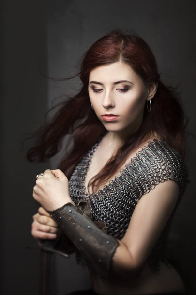 Beautiful girl warrior in chain mail with a sword in his hands. Medieval fantasy valkyrie with long hair posing on a dark background. - Фото, зображення
