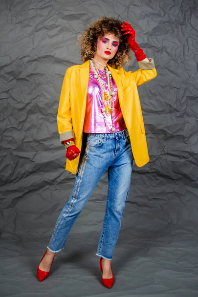 Girl in a yellow jacket and blue jeans with an afro hairstyle. Fashion of the eighties, disco era. Studio photo on gray background. - Fotó, kép