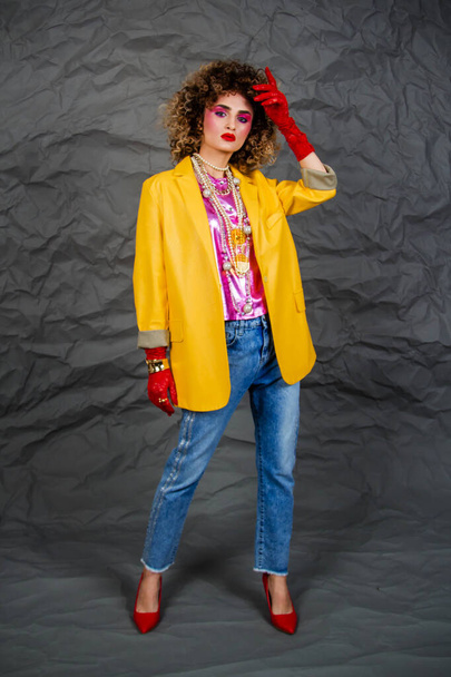 Girl in a yellow jacket and blue jeans with an afro hairstyle. Fashion of the eighties, disco era. Studio photo on gray background. - Zdjęcie, obraz