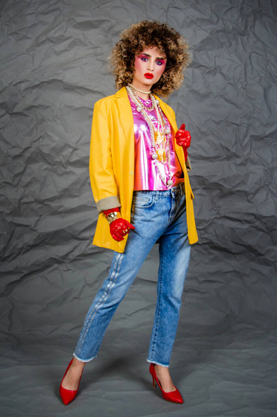 Girl in a yellow jacket and blue jeans with an afro hairstyle. Fashion of the eighties, disco era. Studio photo on gray background. - Valokuva, kuva