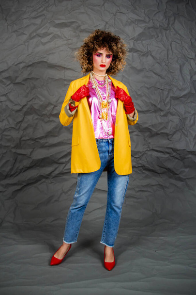 Girl in a yellow jacket and blue jeans with an afro hairstyle. Fashion of the eighties, disco era. Studio photo on gray background. - Foto, afbeelding