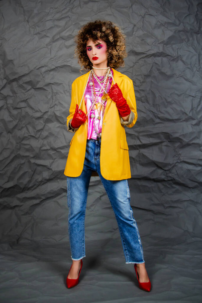 Girl in a yellow jacket and blue jeans with an afro hairstyle. Fashion of the eighties, disco era. Studio photo on gray background. - Photo, image