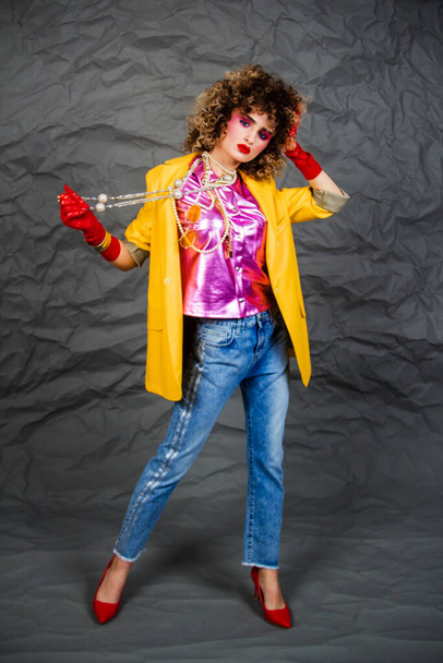Girl in a yellow jacket and blue jeans with an afro hairstyle. Fashion of the eighties, disco era. Studio photo on gray background. - Fotoğraf, Görsel