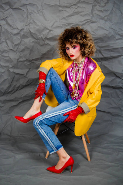 A girl in a yellow jacket and blue jeans with an afro hairstyle sits on a chair. Fashion eighties, the era of disco. Studio photo on a gray background. - 写真・画像