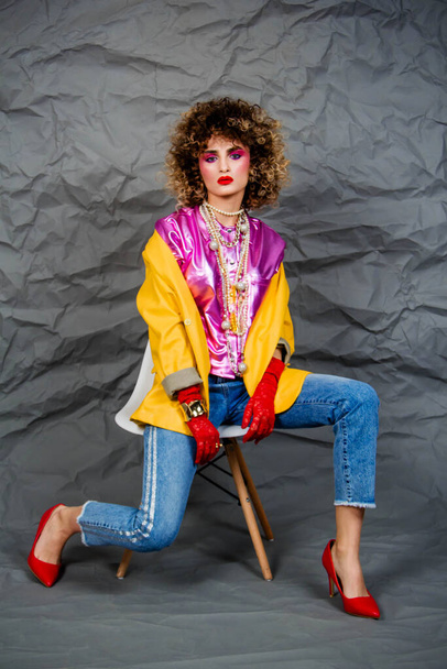 A girl in a yellow jacket and blue jeans with an afro hairstyle sits on a chair. Fashion eighties, the era of disco. Studio photo on a gray background. - Foto, Imagem