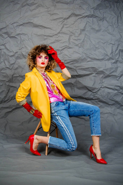 A girl in a yellow jacket and blue jeans with an afro hairstyle sits on a chair. Fashion eighties, the era of disco. Studio photo on a gray background. - Фото, изображение