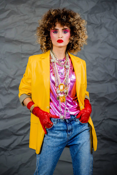 Sarı ceketli ve mavi kotlu, 80 'lerin afro saçlı disko döneminden bir kızın portresi. Stüdyoda gri arkaplanda fotoğraf. - Fotoğraf, Görsel
