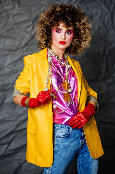 Portrait of a girl in a yellow jacket and blue jeans with afro hair of the eighties, disco era. Photo in studio on a gray background. - Fotoğraf, Görsel