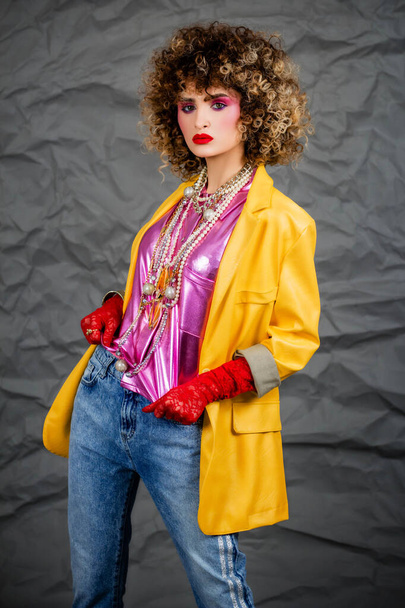 Portrait of a girl in a yellow jacket and blue jeans with afro hair of the eighties, disco era. Photo in studio on a gray background. - Photo, image