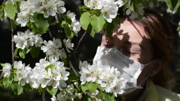 Young woman in medical mask sniffs cherry flowers on a sunny day - Felvétel, videó