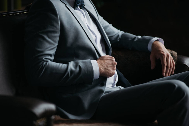 Portrait of a man. The groom in a in a gray suit, white shirt and a bow tie is sitting on a brown leather couch. Room in loft style. - Foto, Imagem