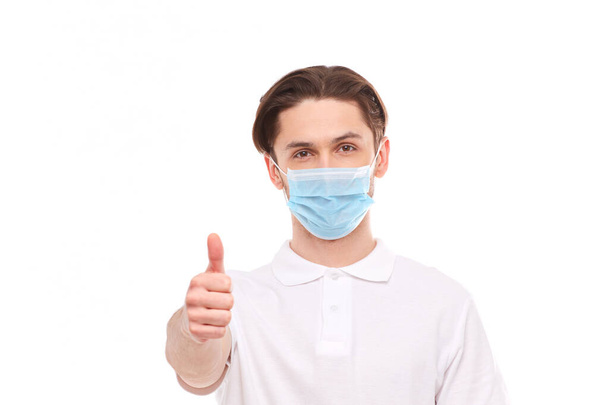 Ein Mann in einem weißen T-Shirt europäischen Aussehens mit medizinischer Maske zeigt den Daumen nach oben. In der Maske sind Sie vor covid 19 Viren geschützt. - Foto, Bild