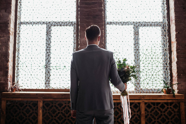 The groom is waiting for the bride in a gray suit in a Moroccan style room. Stylish wedding bouquet in hands. - Foto, afbeelding