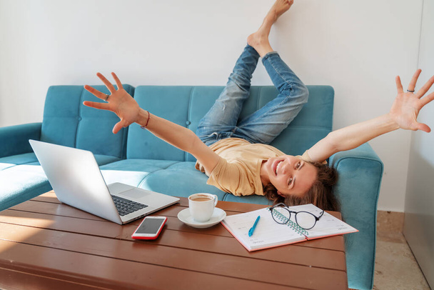 Descanso laboral en casa. mujer atractiva joven emocional se encuentra en un sofá azul frente al lugar de trabajo en casa con un ordenador portátil y una taza de café, feliz y alegre, expresión y emociones
 - Foto, Imagen