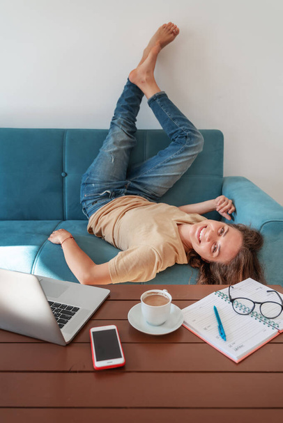 Work break at home. emotional young attractive woman lies on a blue sofa in front of the workplace at home with a laptop and a coffee mug, happy and joyful, expression and emotions - Foto, Imagen