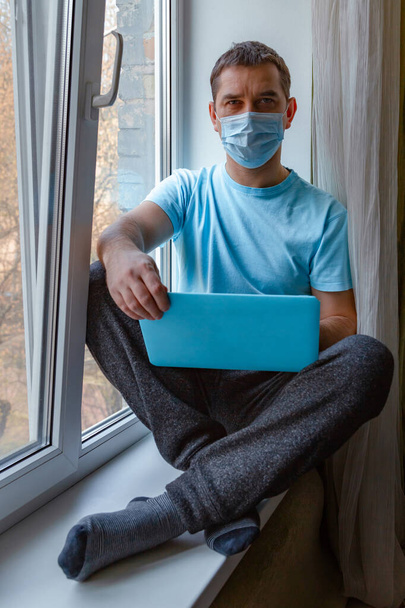 Egy kék orvosi maszkos fiatalember ül egy ablakpárkányon egy laptoppal, és a kamerába néz. Szabadúszó munka, otthoni munka, online tanulás, tanulás, zárlat koncepció. Távolsági oktatás - Fotó, kép