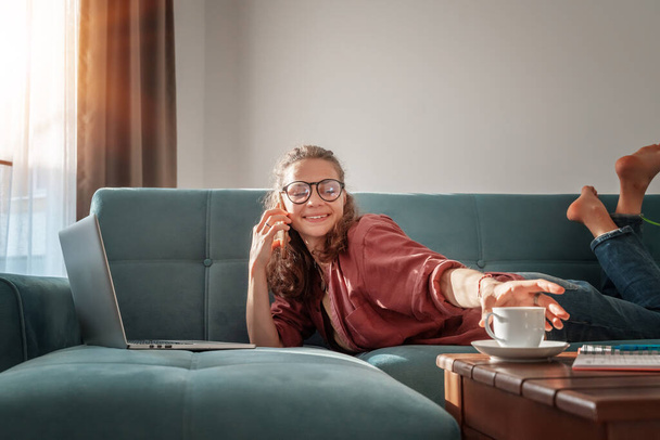 A beautiful young charming girl with glasses lies on her stomach  on a blue sofa in her living room and works on a laptop, distant work, freelancer, work and education online - Фото, изображение