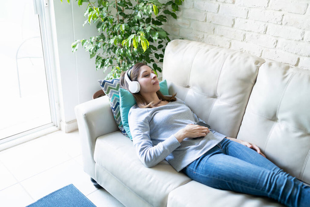 Белая женщина лежит на диване и проводит медитацию дома с наушниками и смартфоном
 - Фото, изображение