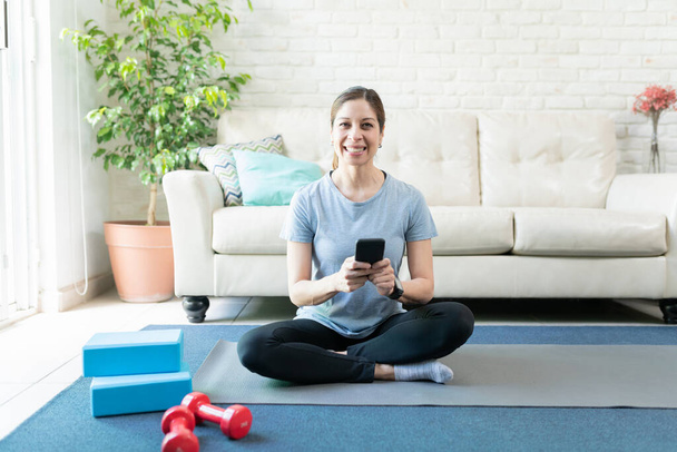Portrét usměvavé aktivní ženy pomocí smartphonu doma při cvičení na podložce jógy - Fotografie, Obrázek
