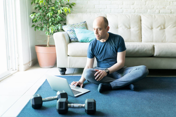 Caucasian bald man looking for some online workouts on a laptop computer and getting ready to exercise and lift some weigths at home - Photo, Image