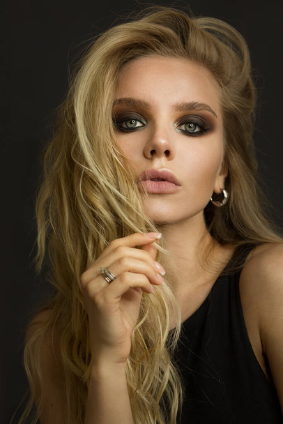 Portrait of a woman model. The blonde holds a lock of blond hair near the face. There is a ring on the finger. Dark makeup smoky eyes. She is looking straight. - Foto, immagini