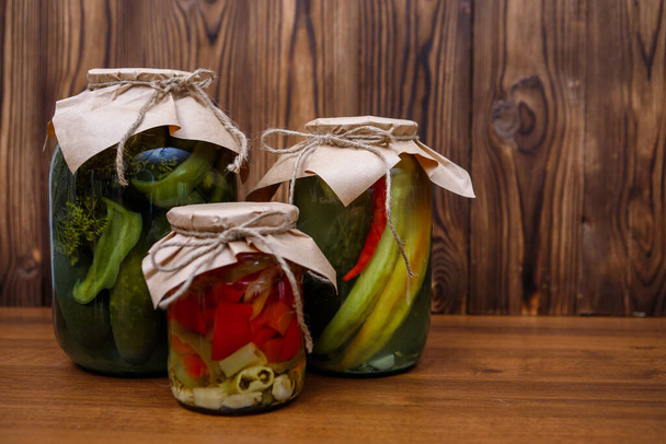 Homemade pickles in glass jars, cucumbers, peppers. Canned vegetables on a wooden background. Preserves. - Фото, зображення
