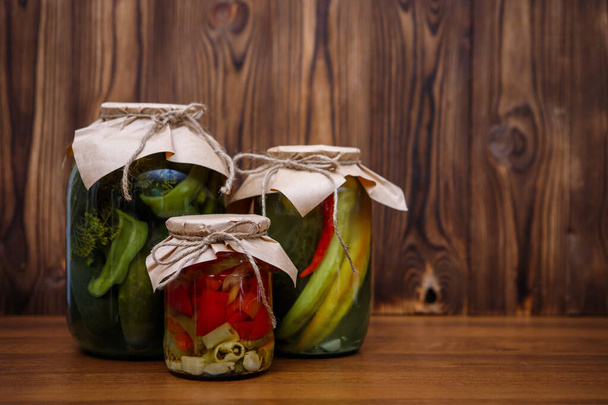 Homemade pickles in glass jars, cucumbers, peppers. Canned vegetables on a wooden background. Preserves. - Фото, изображение