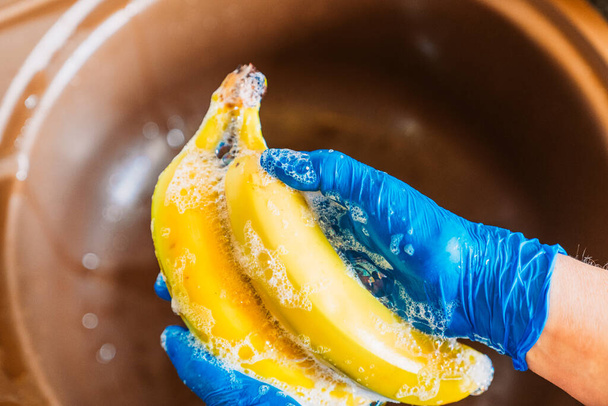 Hände mit blauen Latexhandschuhen desinfizieren Bananen, um die Früchte vom Coronavirus zu dekontaminieren. Obst in der Küchenspüle mit Wasser und Seife waschen, um Viren zu entfernen. - Foto, Bild