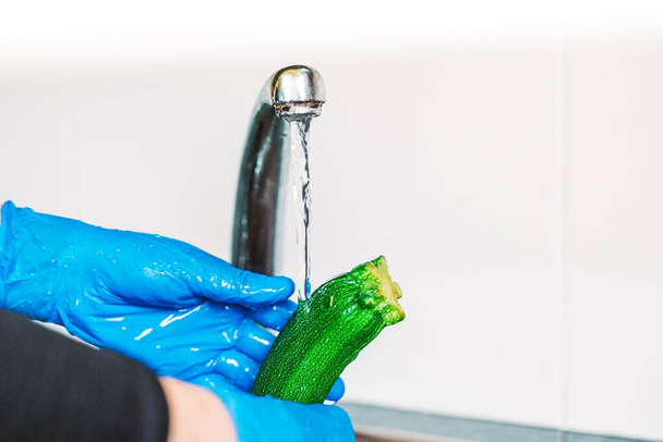 青いラテックス手袋の手は、コロナウイルスから果実を除染するためにズッキーニを消毒します。台所のシンクの野菜を水で洗う. - 写真・画像