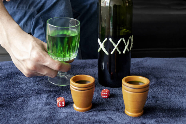 Közel egy férfihoz, aki egy csésze zöld házi likőrt tart két fából készült csészével, zöld kézműves alkoholos üveggel és dupla hat piros kockával. Parti és italos játékok koncepciója - Fotó, kép