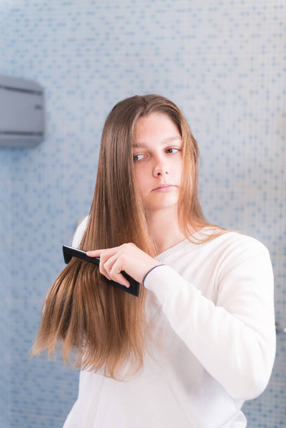 Μια όμορφη ξανθιά κοπέλα χτενίζει τα μαλλιά της μόνη της, κοιτάζεται στον καθρέφτη στο μπάνιο, πρωινή ρουτίνα, επιλεκτική εστίαση. - Φωτογραφία, εικόνα