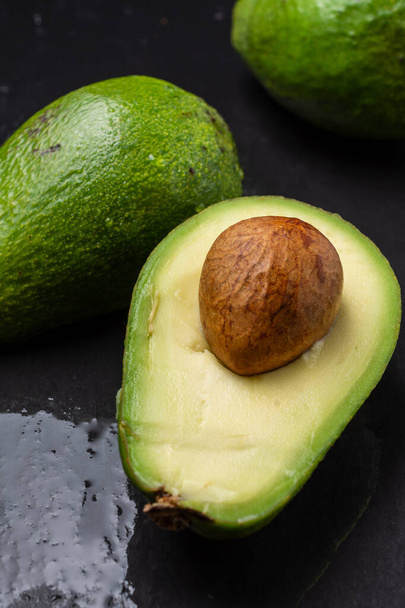 Вид з повітря на зелені авокадо, один вирізаний з насінням, на вологому чорному шифері, вертикальний
 - Фото, зображення