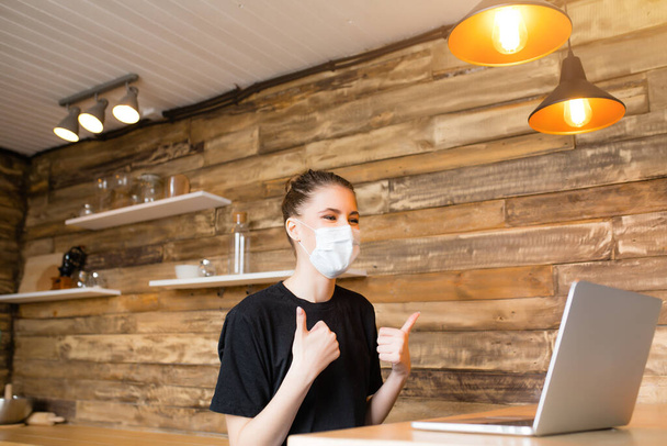 Girl in kitchen with laptop shows the gesture "thumbs up" and smiling through the medical mask - Foto, afbeelding