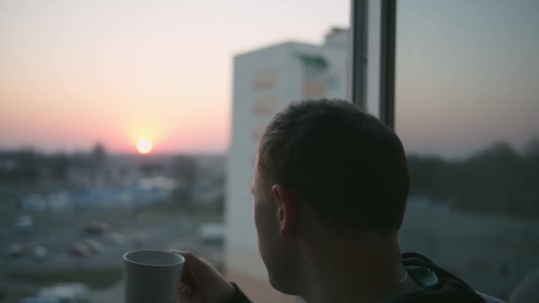 Facet z kubkiem herbaty stoi na balkonie podczas zachodu słońca. - Materiał filmowy, wideo