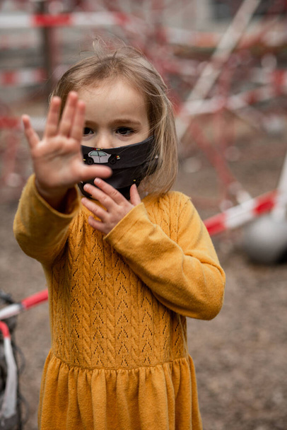 Cute little girl noszenie maski i stojąc w pobliżu zamkniętego placu zabaw podczas blokady koronawirusa - Zdjęcie, obraz