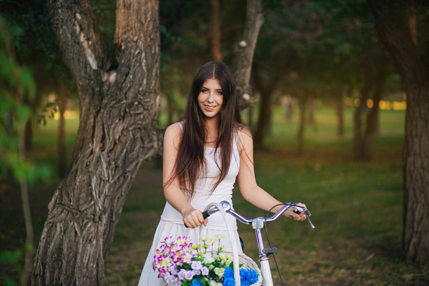 Portrait of a girl in a white T-shirt and skirt with a bicycle and a basket of flowers in the park in the evening. - Foto, Imagem