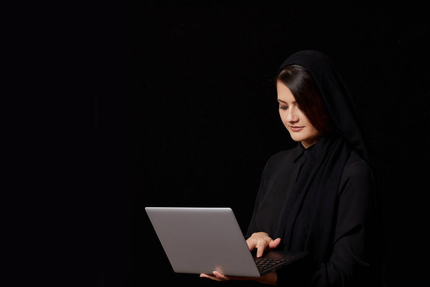 a young brown-haired woman in black clothes with a covered head with a laptop in her hands. on a black isolated background. - Valokuva, kuva