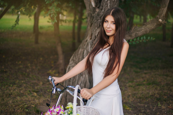 Portrait of a girl in a white T-shirt and skirt with a bicycle and a basket of flowers in the park in the evening. - Fotoğraf, Görsel