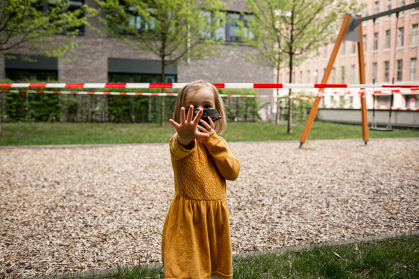 Cute little girl noszenie maski i stojąc w pobliżu zamkniętego placu zabaw podczas blokady koronawirusa - Zdjęcie, obraz