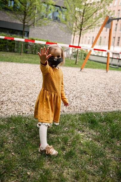 Linda niña usando una máscara y de pie cerca de un patio cerrado durante el bloqueo coronavirus
 - Foto, Imagen