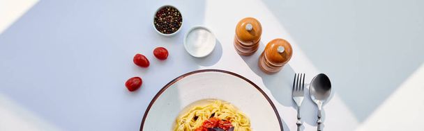 верхний вид вкусной пасты с помидорами подается со столовыми приборами, соль и перец мельницы на белый стол при солнечном свете, панорамный урожай
 - Фото, изображение