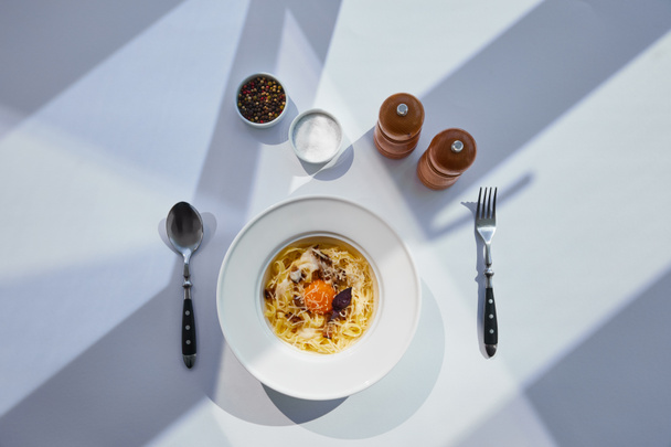 Blick von oben auf köstliche Pasta Carbonara serviert mit Besteck, Salz- und Pfeffermühlen auf weißem Tisch im Sonnenlicht - Foto, Bild