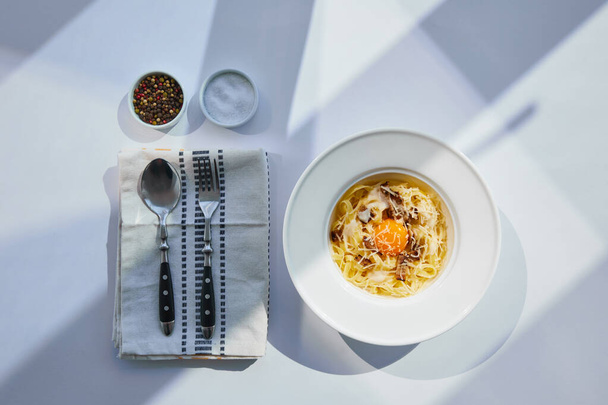 vista superior de la deliciosa pasta carbonara servida con cubiertos y condimentos en mesa blanca a la luz del sol
 - Foto, Imagen