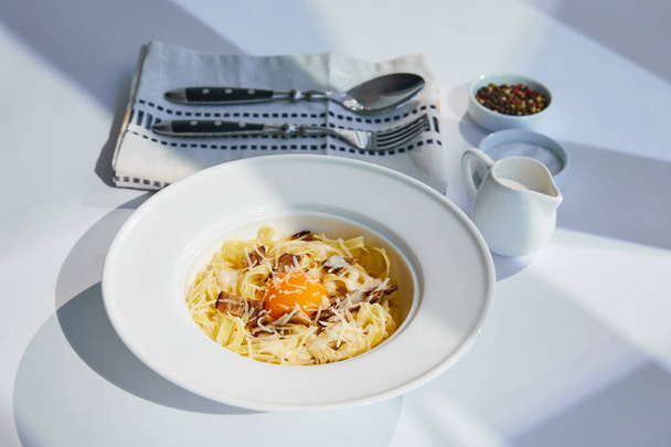 delicious pasta carbonara served with cutlery, cream and seasoning on white table in sunlight - Foto, imagen