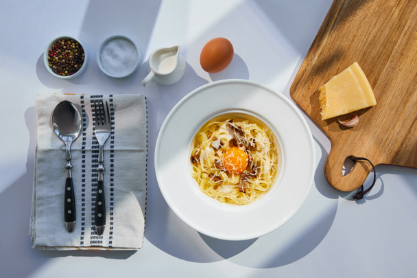 Blick von oben auf köstliche Pasta Carbonara serviert mit Besteck, Gewürzen und Zutaten auf weißem Tisch im Sonnenlicht - Foto, Bild