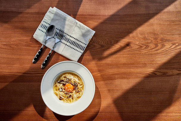 Blick von oben auf köstliche Pasta Carbonara serviert mit Besteck und Serviette auf Holztisch im Sonnenlicht - Foto, Bild