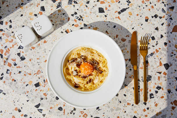 верхний вид вкусной пасты карбонара подается с золотыми столовыми приборами, соль и перцовые шейкеры на каменном столе в солнечном свете
 - Фото, изображение