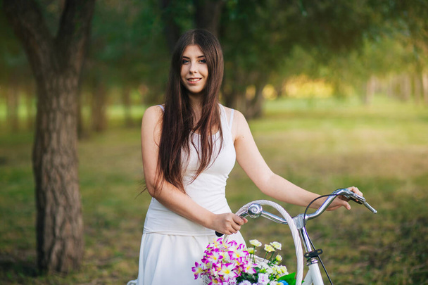  Joven mujer sonriente se sienta en una bicicleta con una cesta de flores en la noche en el parque
. - Foto, Imagen